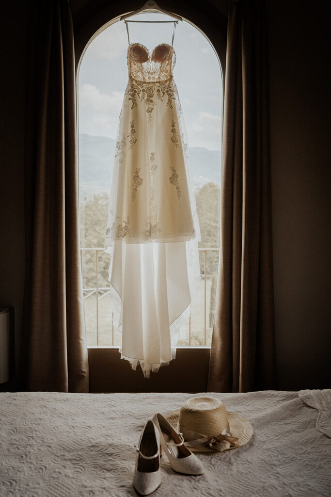 Aufnahme des Brautkleides hängend am Fenster mit den Brautschuhen auf dem Bett in Szene gesetzt beim Getting Ready im Zimmer der Braut
